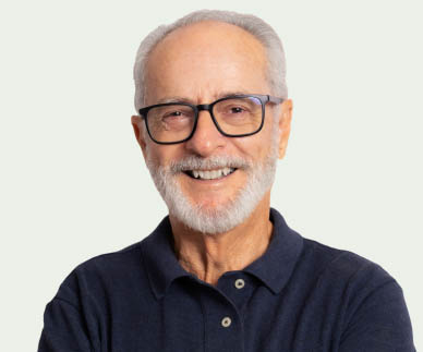 A mature man smiling. He has white facial hair, is wearing glasses and a dark blue polo shirt