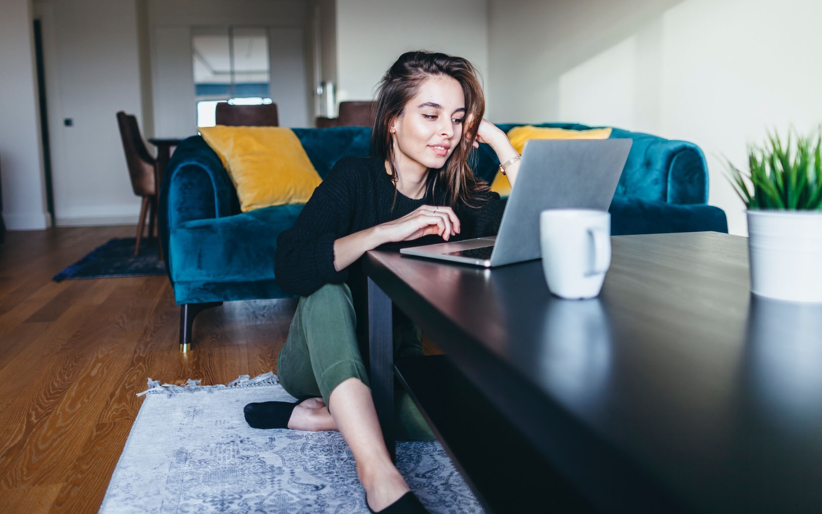 A woman sits on the living room floor while looking at her laptos