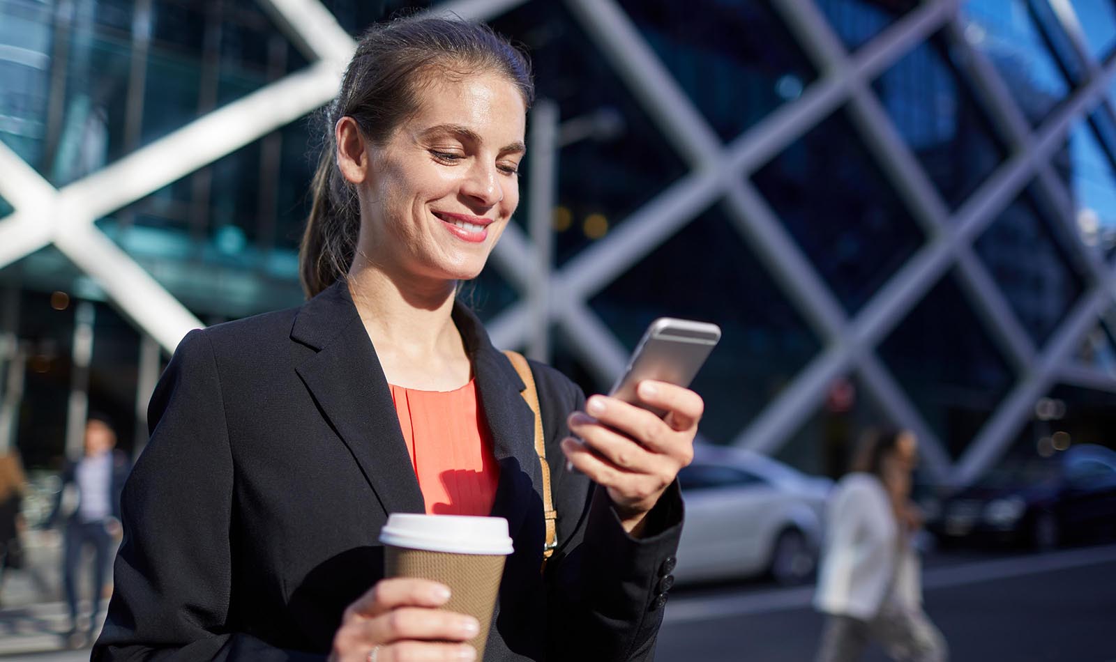 A smiling woman looks down at her phone will standing outside a building in a business district